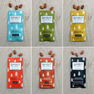 SPIRIT Almond Japanese Style Dry Roasted Almonds colorful lineup, six flavors.  Koji Salt, light blue. Seaweed, green. Mustard, yellow. Curry, reddish orange. Miso, brownish-red. Black Garlic, black. Each in 1.12 oz. single serving bags and a few almonds of each flavor above each bag.